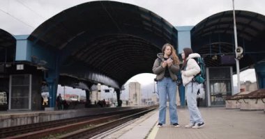 Sırt çantalı iki genç kadın tren istasyonunda selfie çekiyor. Yolculukları için heyecanlılar ve anı yakalamak için sabırsızlanıyorlar..