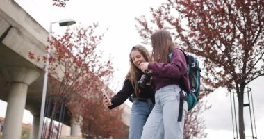 Sırt çantalı ve kameralı iki genç kadın küçük bir çiçeğe bakıyor ve şehir ortamında fotoğraf çekiyor..