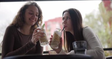 İki genç kadın açık bir kafede buzlu kahve içip sohbet ediyorlar. Gülüyorlar ve bağlanıyorlar, anın tadını çıkarıyorlar..