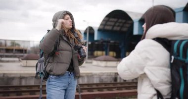 Tren istasyonunda sırt çantalı iki genç kadın, biri diğerinin fotoğrafını çekiyor. Seyahat maceralarının tadını çıkarıyorlar..