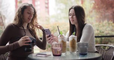 İki genç kadın selfie çekiyor ve dışarıda bir kafede eğleniyorlar. Gülümsüyorlar, gülüyorlar, içkilerinin tadını çıkarıyorlar ve birbirlerine eşlik ediyorlar..