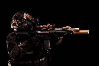 Gece görüş gözlükleri ve kamuflaj kıyafetleriyle donatılmış bir taktik asker tüfeğini siyah bir ayara doğrultuyor. Sahnede hazırlık, ihtiyat ve askeri profesyonellik duygusu aktarılıyor.