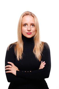 Profesyonel bir iş kadını kollarını kavuşturarak güvenle ayakta durur. Uzun sarı saçları var ve siyah bir balıkçı yaka giyiyor. Güçlü bir tavır ve odaklanmış bir ifade sergiliyor..