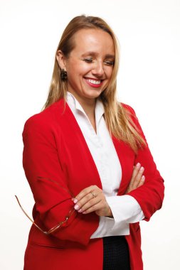 Kendine güvenen bir iş kadını elinde okuma gözlüğü tutarken gülümseyen parlak kırmızı bir ceket ve beyaz bir bluz giyer. Temiz bir stüdyo ortamında canlı ve profesyonel bir imaj çiziyor..