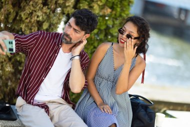 Genç bir çift, güneşli bir günün tadını çıkarırken suyun kenarında selfie çekiyor. Sıradan bir gezinti sırasında keyifli bir anı paylaşıyorlar..