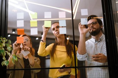 Meslektaşlar parlak bir ofisin cam duvarındaki renkli yapışkan notları kullanarak beyin fırtınası yapıyorlar. Fikirlerini ifade ederler ve bu yaratıcı ortamda bir projede işbirliği yaparlar..