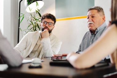 İki işadamı modern bir ofis alanında derin bir sohbetin içindeler. Diğerleri dikkatle dinlerken, onlar belgeleri analiz edip stratejik planlamaya giriyorlar. Atmosfer işbirlikçi..