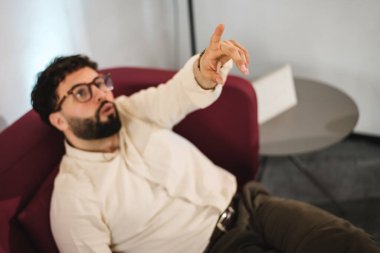 Bir işadamı çağdaş bir kanepede otururken bir sunum yapıyor, sanki bir noktaya değiniyormuş gibi el kol hareketi yapıyor. Kurulumda, işbirliği öneren şık bir ofis ortamı yer alıyor.