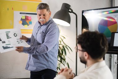 Bir işadamı modern bir ofisteki özenli bir meslektaşına veri analiz çizelgeleri sunar. Ayarda tartışmayı geliştiren parlak ışıklar ve canlı görseller yer alıyor.