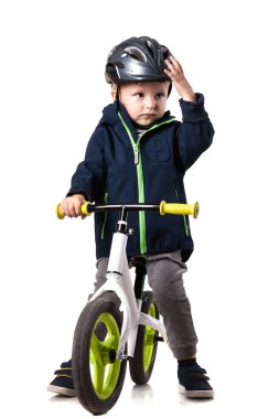 Küçük bir çocuk, yeşil tekerlekli beyaz bir bisiklette, kask takarak dengesini sağlarken kararlılık gösteriyor. İçeride duruyor, heyecan verici bir yolculuğa hazır..