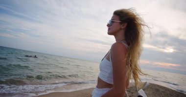 Genç bir kadın sahilde duruyor, saçları rüzgarda dalgalanırken okyanus dalgalarına hayranlık duyuyor. Gün batımı güzel bir parıltı yayar, huzurlu bir atmosfer yaratır..