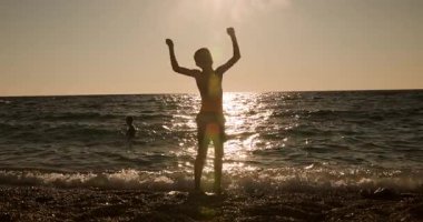 A striking silhouette of a young child celebrating freedom and hope on a tranquil beach as the sun sets over the glistening ocean waves, hands raised in triumph.