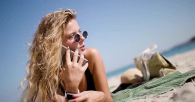 Bir kadın güzel bir kumsalda, telefonu kulağına dayalı bir havluyla dinleniyor. Güneşlenirken selfie çeker ve dalgaların seslerinden zevk alır. Eğlenceli bir anı yaratır..