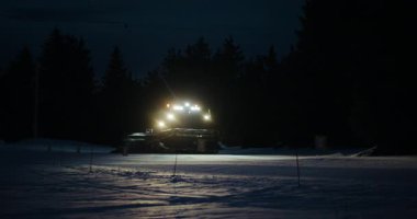 Bir kar kedisi geceleri bir kayak yamacını tımar eder. Kar arabalarının ışıkları karları ve etrafındaki ağaçları aydınlatıyor..