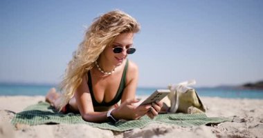 Kumsalda uzanmış, kumsalda uzanan bir kadın telefonda konuşurken gülümsüyor. Arka planda okyanus parıldıyor, barışçıl ve neşeli bir atmosfer yaratıyor..