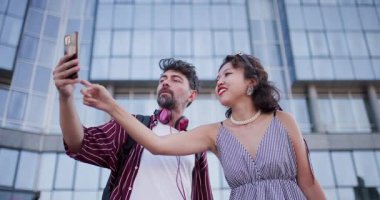 Bir çift modern cam bir binanın önünde durup selfie çekiyor, parlak bir günde şehir yaşam tarzını, bağlantıyı ve mutluluğu simgeliyor..