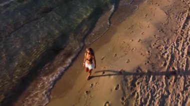 Kadın güzel bir kumsalın kıyısında yürüyor dalgalar kumu sararken ayak izleri bırakıyor, huzur verici titreşimler yaratıyor..