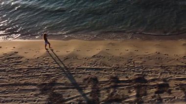 Kadın gün batımında plaj boyunca koşar. Atletik figürünün ve kıyıya çarpan dalgaların havadan görünüşü..