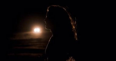 Bir kız gecenin karanlığında ıssız bir yolda, arabanın arka planındaki farlara karşı gölgede duruyor. Karanlık bir ortamda entrika ve yalnızlık hissi yaratıyor..