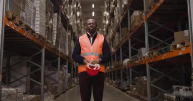 Güvenlik donanımlı bir depo işçisi, turuncu yelek ve kırmızı miğfer dahil, raflar ve kutularla çevrili büyük bir depo tesisinde güvenle duruyor..