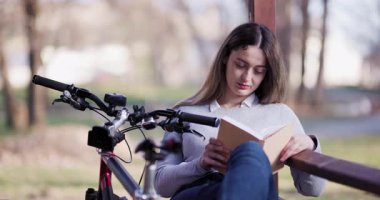Huzurlu bir parkta yakalanan bu resim, yanında bisikletiyle kitap okuyan, dışarıda huzurlu bir anın tadını çıkaran genç bir kadını gösteriyor..