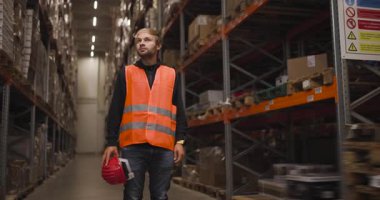 Turuncu güvenlik yeleği ve kırmızı başlık giyen bir depo işçisi raflar ve kutularla dolu büyük bir endüstriyel depolama tesisinde dikilirken yukarı bakıyor..