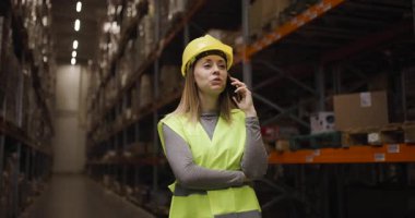 Güvenli yelek ve kask giyen kendine güvenen depo çalışanı, envanter dolu raflarla çevriliyken telefonla iletişim kuruyor. Endüstriyel lojistikteki verimliliği ve organizasyonu vurguluyor..