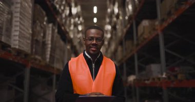 Depo çalışanı, envanter kontrolü yaparken gülümsüyor. Koridorlar düzgünce istiflenmiş paketlerle dolu, yoğun bir depolama tesisinde organizasyon ve endüstriyel verimliliği vurguluyorlar..