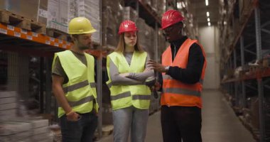 Çelik yelek ve kask giyen üç depo çalışanı, yoğun bir depolama ortamında ekip çalışması ve güvenliğini ön plana çıkaran bir tarayıcı kullanarak sohbete girişiyor..