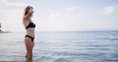 Genç bir kadın plajın sığ sularında geziniyor, güneş ışığını emiyor. Siyah bir bikini ve güneş gözlüğü takıyor. Okyanusun rahat bir yaz havasını somutlaştırıyor..