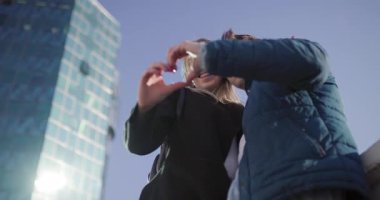 Genç, neşeli bir çift modern bir binanın önünde duruyor. Elleriyle bir kalp şekli oluşturuyorlar. Şehir ortamında aşkı ve birlikteliği simgeliyorlar..