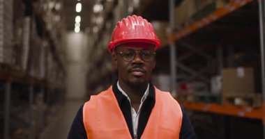 Dolu bir depoda duran çelik yelek ve miğferli bir depo çalışanının portresi. Güvenlik, profesyonellik ve endüstriyel çalışma ortamını temsil eder.