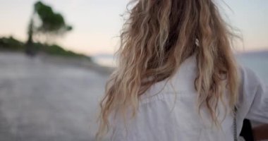 Kıvırcık saçlı bir kadın sakin bir plajda yürüyor ve büyüleyici günbatımı manzarasını seyrediyor. Gün sona ererken arka planda sohbet eden ve gülen arkadaşlar görülüyor..