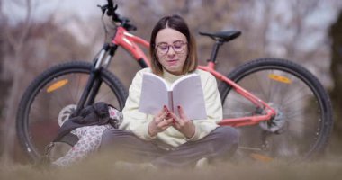 Açık havada kitap okumaya dalmış, turuncu dağ bisikleti yanında park etmiş genç bir kadını gösteren sakin bir sahne..