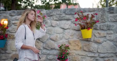 Güzel bir kadın kırsal taştan bir duvarda zarif bir şekilde yürüyor, etrafı saksılarda canlı çiçeklerle çevrili. Gündüz güneşinde huzurlu bir atmosfer yakalıyor ve onun zarafetini arttırıyor..