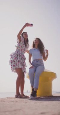Sahil rıhtımında selfie çeken iki genç kadının berrak gökyüzünün altında neşeli bir anı paylaştığı canlı bir sahne..