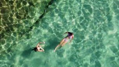 Güneşli bir günde kristal berrak turkuaz sularda yüzmeyi seven iki kadın. Yaz tatilindeki dostların hava aracı görüntüsü.