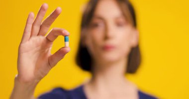 Sağlığını ve sağlığını vurgulayan, sarı arka planda mavi ve beyaz kapsül hapı sunan gülümseyen genç bir kadının yakın çekimi..