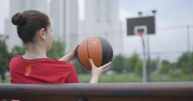 Genç bir bayan basketbol oyuncusu, şehir merkezindeki bir bankta otururken basketbol topunu çeviriyor. Yetenek ve konsantrasyonu somutlaştırıyor..
