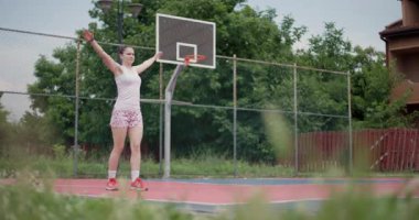 Bayan basketbolcu dışarıda sahada esneyerek antrenmana hazırlanıyor. Sahne, çevresi yeşilliklerle çevrili bir yerleşim bölgesinde atletik bir kadını yakalıyor..