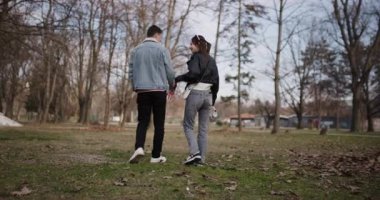 Genç bir çift parkta yürüyor, el ele tutuşuyor ve güzel bir günde birbirlerine eşlik ederek eğleniyorlar..