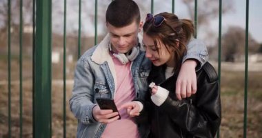 Genç bir çift parkta birlikte vakit geçirir, telefonunu paylaşır ve gülümser..