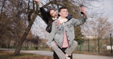 Genç bir çift parkta selfie çekiyor. Kız, oğlanların sırtında gülümseyip kameraya el sallıyor..