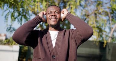 Mutlu bir genç adam bir park ortamında kulaklıkla müzik dinlemekten hoşlanır. Gülümsüyor ve müziğe el sallıyor..