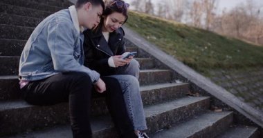 Genç bir çift parkta basamaklarda oturuyor, birlikte akıllı telefona bakıyor ve gülümsüyor. Günlük giysiler giymişler..