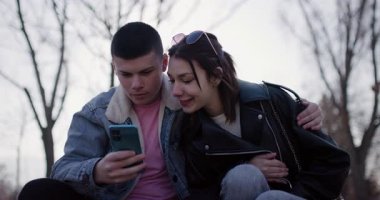 Genç bir çift dışarıda merdivenlerde birlikte oturur, sarılırlar ve akıllı telefona bakarlar. Gülümsüyorlar ve mutlular.