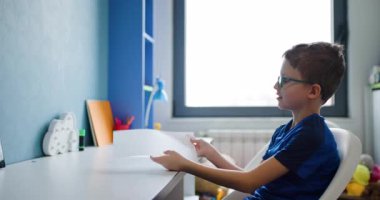 Genç bir çocuk masasında oturuyor ve bir parça kağıt katlıyor. O, gözlük ve mavi bir gömlek giyiyor..