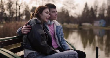 Genç bir çift gölün kenarındaki bankta oturuyor, kulaklıklarını paylaşıyor ve birlikte müzik dinliyorlar..