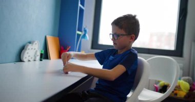 Gözlüklü genç bir çocuk odasında masasında oturuyor ve kağıt uçak yapıyor..