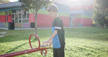 Genç bir çocuk oyun parkında tahterevalli üzerinde duruyor, tahterevalli yüzeyine küçük taşlar ya da tohumlar bırakıyor..